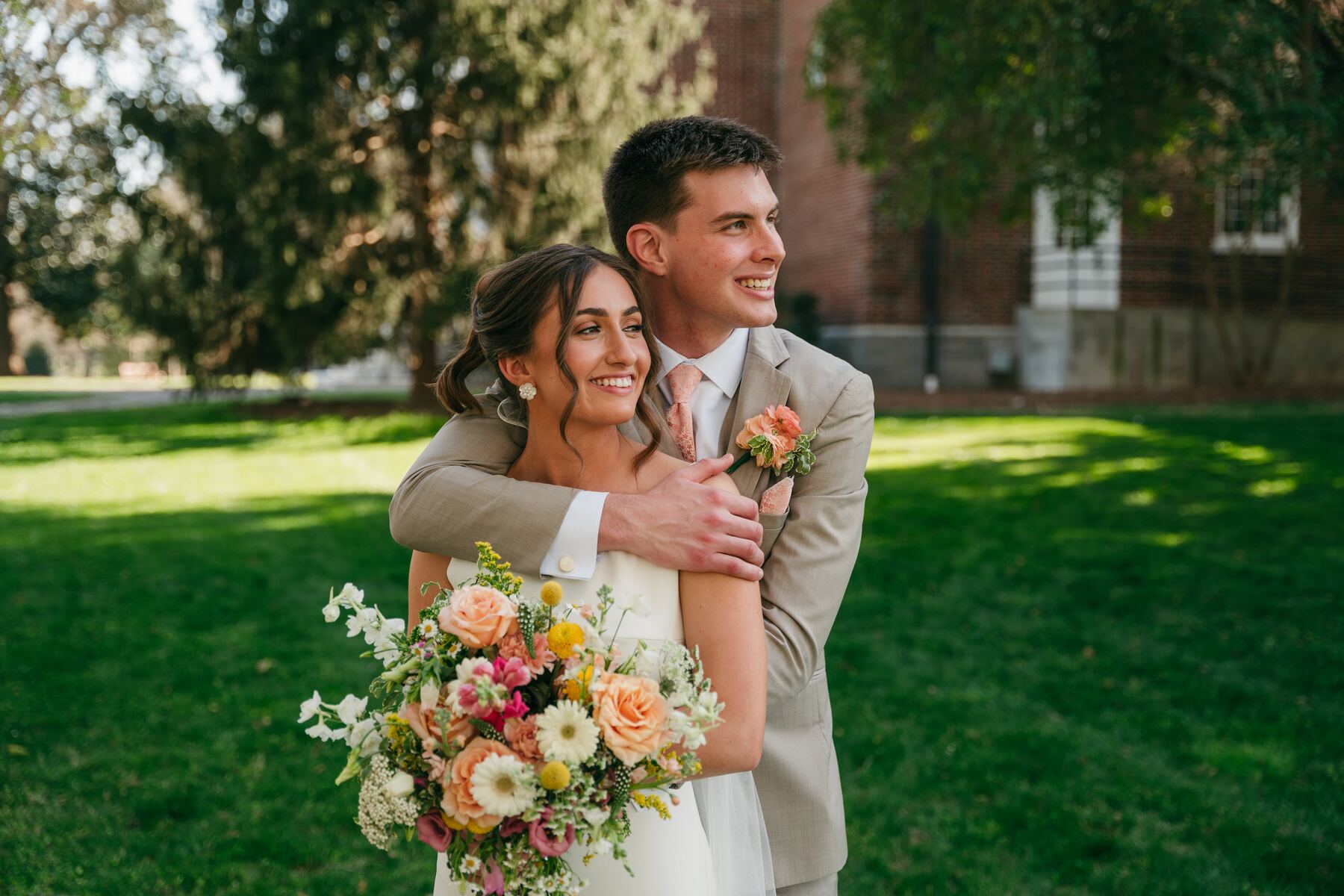 A wide frame of the couple embracing on the lawn, bouquet in hand