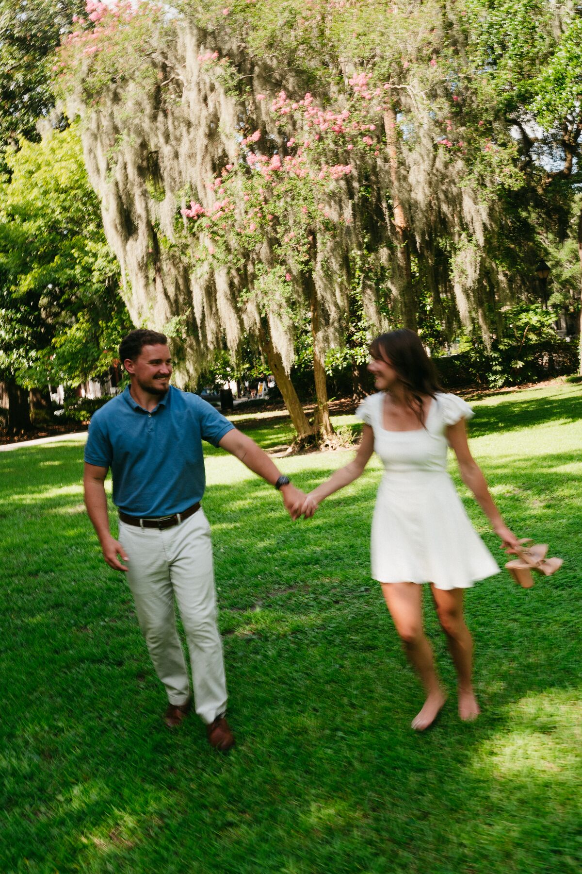 An engaged couple walking hand in hand beneath Spanish moss in Forsyth Park, Savannah